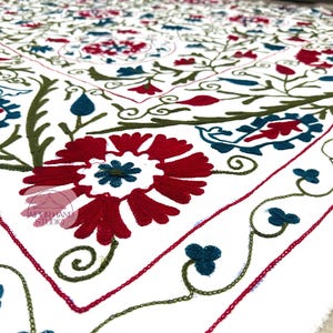 Puede incluir: Textil blanco con intrincados bordados. El diseño presenta patrones florales rojos, turquesa y verdes. Grandes flores rojas con centros turquesa están rodeadas de vides verdes y acentos turquesa.