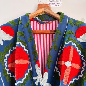 May include: A blue embroidered jacket with a pink and white striped lining. The embroidery features red, white, and green flowers.