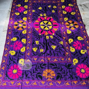 May include: A vibrant purple textile with intricate floral embroidery in pink, yellow, and orange. The design features large central flowers and smaller floral motifs, framed by a decorative border. The text "JAIPUR HAND STUDIO" is visible.