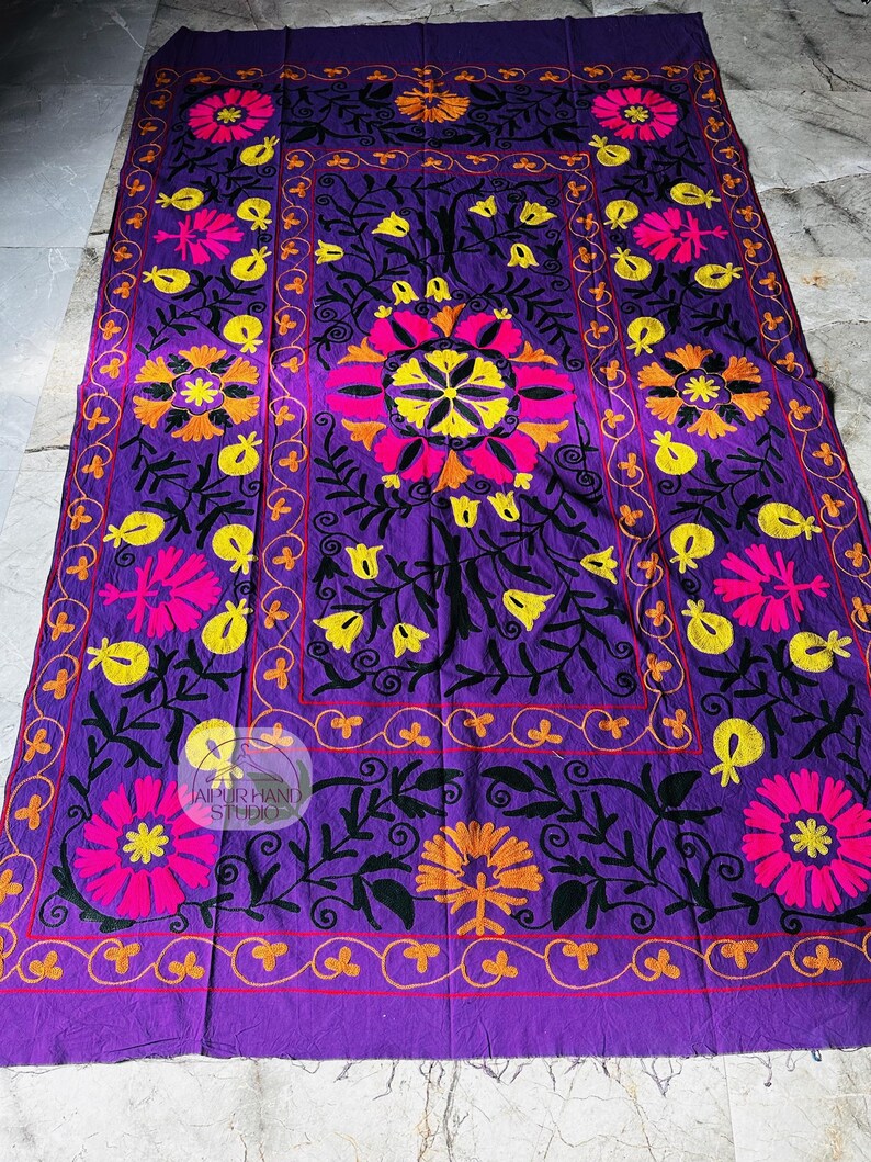 May include: A vibrant purple textile with intricate floral embroidery in pink, yellow, and orange. The design features large central flowers and smaller floral motifs, framed by a decorative border. The text "JAIPUR HAND STUDIO" is visible.