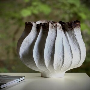 May include: A white, textured ceramic vase with a unique, fluted design. The vase has a bulbous shape with vertical ridges and a dark brown rim. The vase sits on a white surface, with a book visible in the foreground.
