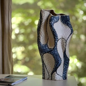 May include: A tall, textured ceramic vase with a unique shape. The vase features a white base with blue, organic-shaped patterns outlined in black. The vase is displayed on a white surface near a window with a blurred green background.