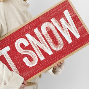 Close up of the barn red "Let It Snow" sign held in hand, showing the distressed white lettering texture.