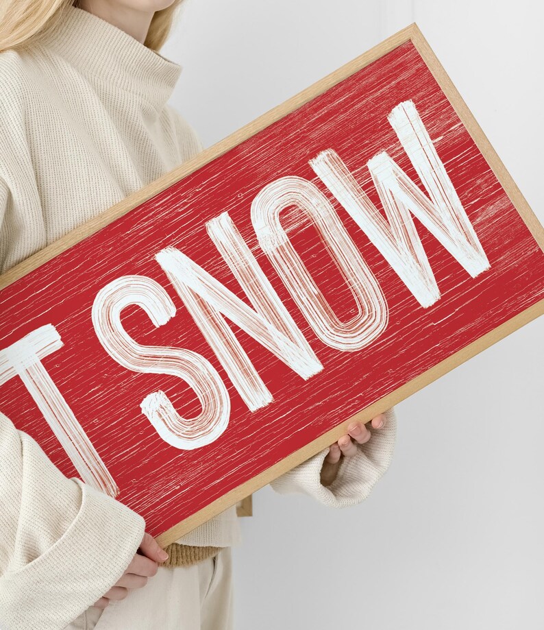 Close up of the barn red "Let It Snow" sign held in hand, showing the distressed white lettering texture.