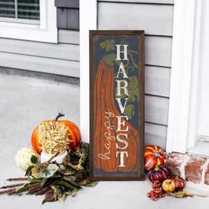 Tall harvest sign in navy leaning on white siding with pumpkins and a harvest bouquet.