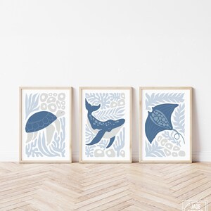 A dusty blue nursery sea creature print trio leans on a white wall above a light herringbone wood floor.