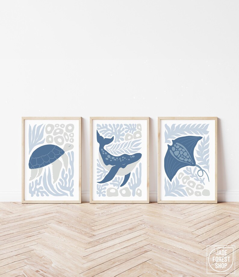 A dusty blue nursery sea creature print trio leans on a white wall above a light herringbone wood floor.
