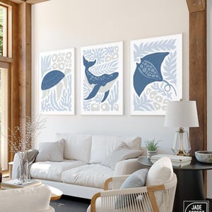 A sea creature print set of a turtle, a whale and a ray in dusty blue hangs under a wood beam above a white sofa.