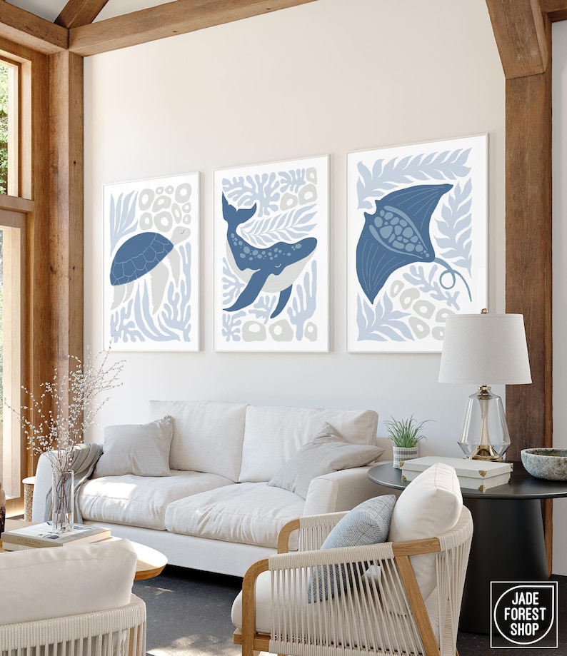 A sea creature print set of a turtle, a whale and a ray in dusty blue hangs under a wood beam above a white sofa.