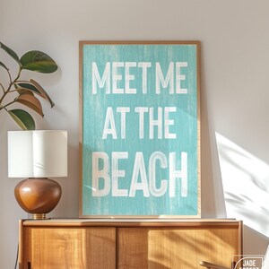 The teal Meet Me At The Beach sign in a wood frame leaning on a wood console with a lamp and a plant.