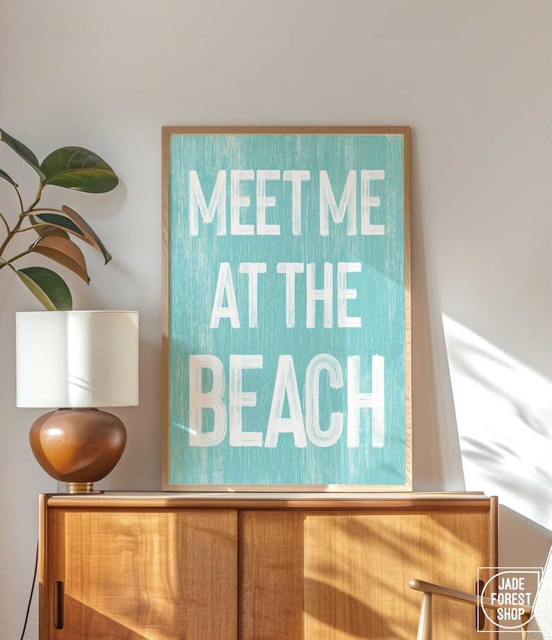 The teal Meet Me At The Beach sign in a wood frame leaning on a wood console with a lamp and a plant.
