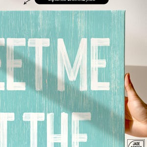 A hand touching the corner of the Meet Me At The Beach sign, showing the lettering up close as the unframed canvas print option.