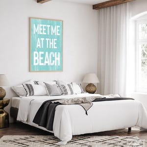 The teal Meet Me At The Beach sign in a wood frame displayed on a white wall above a bed with pillows.