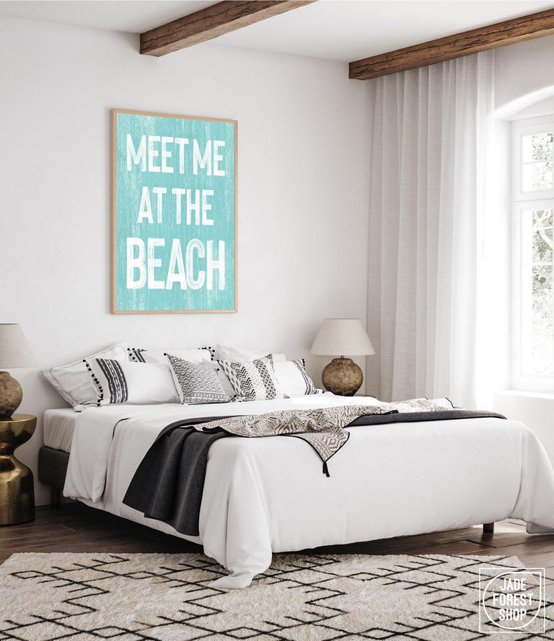The teal Meet Me At The Beach sign in a wood frame displayed on a white wall above a bed with pillows.