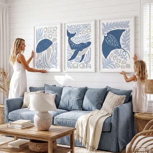 A print set of a sea turtle, a whale and a stingray is hung by a woman and a young girl above a blue sofa.