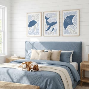 A dusty blue sea creature print set hangs above a bed with a blue quilt, a sleeping dog and a tufted headboard.