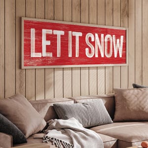"Let It Snow" farmhouse sign in barn red, displayed on a wood paneled wall above a tan sofa.