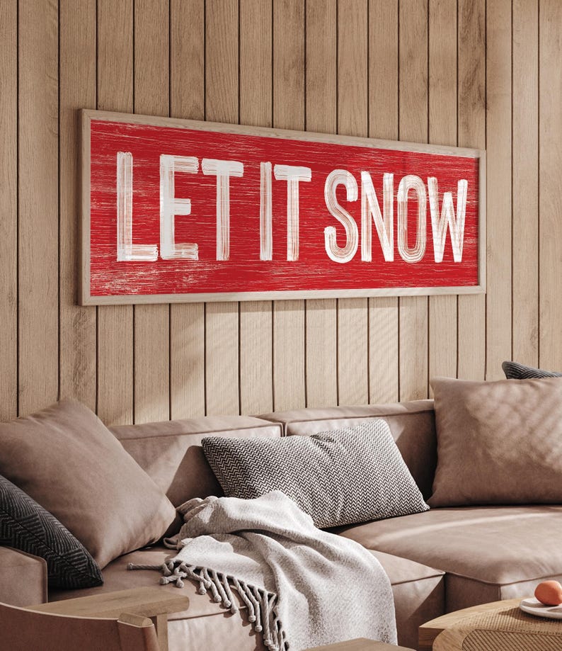 "Let It Snow" farmhouse sign in barn red, displayed on a wood paneled wall above a tan sofa.