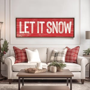 Barn red "Let It Snow" sign with weathered white lettering in a black frame, hung above a white sofa with plaid pillows.