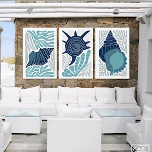 May include: Three framed art prints featuring stylized seashell designs in shades of blue and teal against a white background. The prints are displayed on a stone wall above white outdoor furniture, creating a coastal theme.