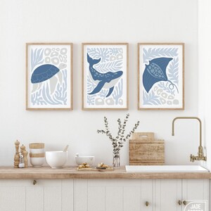 A sea creature design print set in dusty blue hangs above a kitchen counter with white cabinets, a brass faucet and a board.