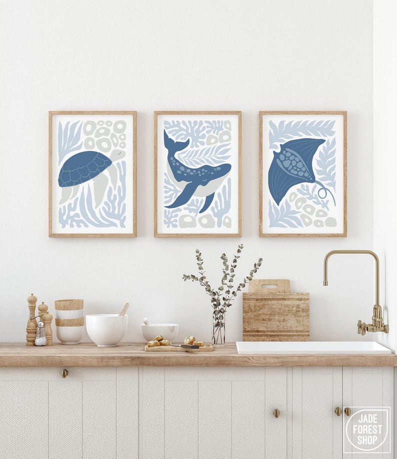 A sea creature design print set in dusty blue hangs above a kitchen counter with white cabinets, a brass faucet and a board.
