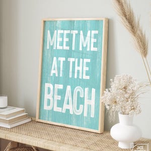 The teal Meet Me At The Beach sign in a wood frame leaning on a wood table with books and a vase.