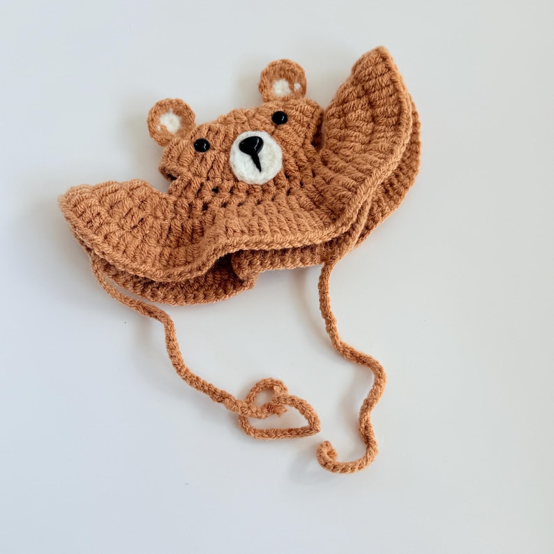 May include: A brown crocheted bear hat with a white face and black eyes. The hat has a long brown crocheted tie.