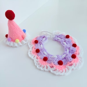 May include: A crocheted party hat and collar set. The hat is pink with a red pom-pom and colourful pom-poms around the base. The collar is a layered design of pink, purple, and white with red pom-poms.
