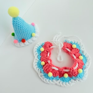 May include: A crocheted party hat and bib set. The hat is light blue with a yellow pom-pom and colourful pom-pom accents. The bib is red, white, and blue with yellow pom-pom accents. The set is handmade and suitable for a pet.