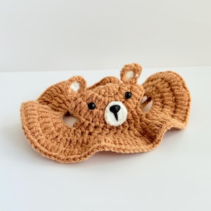 May include: A brown crocheted bear head with black eyes and a white nose. The bear head is attached to a brown crocheted blanket with a ruffled edge.