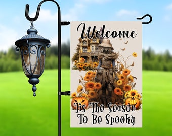 Pode incluir: Uma bandeira de jardim com tema de Halloween. A bandeira apresenta uma casa assombrada, girassóis, um espantalho e abóboras. O texto na bandeira diz "Welcome" e "Tis The Season To Be Spooky". Está pendurada em um poste de jardim de metal preto com uma lanterna decorativa.