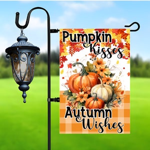 May include: A fall-themed garden flag with a white background and orange and brown checkered pattern at the bottom. The top portion features a watercolor illustration of pumpkins, fall leaves, and flowers. The text "Pumpkin Kisses Autumn Wishes" is written in black lettering.