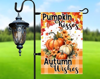 Op de afbeelding: Een herfst-thema tuin vlag met een witte achtergrond en een oranje en bruin geruit patroon aan de onderkant. Het bovenste gedeelte heeft een aquarel illustratie van pompoenen, herfstbladeren en bloemen. De tekst "Pumpkin Kisses Autumn Wishes" is geschreven in zwarte letters.