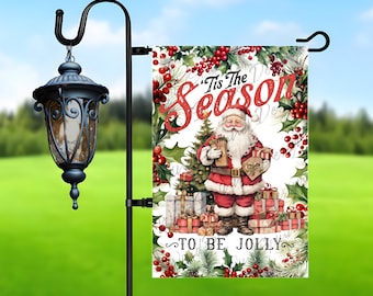 Pode incluir: Uma bandeira de jardim com tema natalino com fundo branco e uma borda de coroa vermelha e verde. O centro da bandeira apresenta um Papai Noel de desenho animado segurando presentes, uma árvore de Natal e o texto "Tis the Season to Be Jolly".