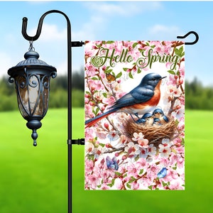 Puede incluir: Una bandera de jardín con una familia de azulejos en un nido rodeado de flores rosas y mariposas. La bandera dice "Hello Spring". Está colgada de un poste de jardín de metal negro con una linterna decorativa.
