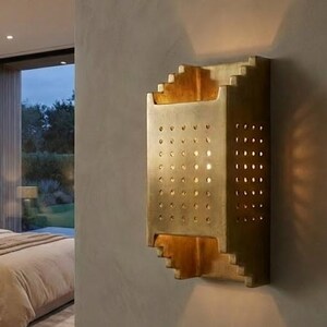 Könnte beinhalten: Goldfarbene Wandleuchte mit geometrischem Design und mehreren kleinen kreisförmigen Perforationen. Die Leuchte ist an einer neutralfarbenen Wand montiert und wirft einen warmen Schein. Die Schlafzimmereinrichtung umfasst ein Bett mit neutralfarbener Bettwäsche.