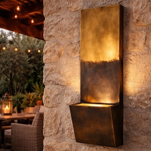 May include: A bronze outdoor wall light is mounted on a stone wall. The light has a rectangular design with a long, vertical section and a downward-facing light. The light is part of an outdoor lighting fixture.
