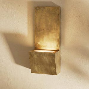 May include: A brass wall sconce featuring a rectangular backplate and a cube-shaped light fixture. The sconce emits a warm light. The fixture is mounted on a textured, off-white wall, creating a contrast between the smooth metal and the rough surface.