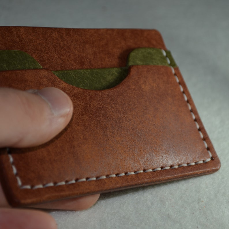 May include: A brown leather card holder with olive green interior pockets. The edges are stitched with white thread. The card holder is rectangular and appears to be handmade.