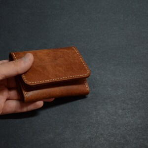 以下が含まれることがあります： 白いステッチが付いた茶色の革財布。財布は閉じており、手で持たれています。