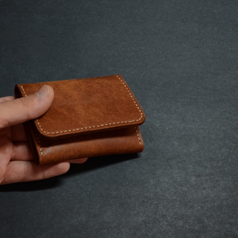 以下が含まれることがあります： 白いステッチが付いた茶色の革財布。財布は閉じており、手で持たれています。