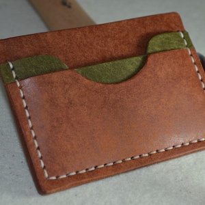 May include: A handmade leather card wallet in shades of brown and olive green. The wallet features a contrasting olive green pocket and is stitched with white thread. The leather has a textured appearance.
