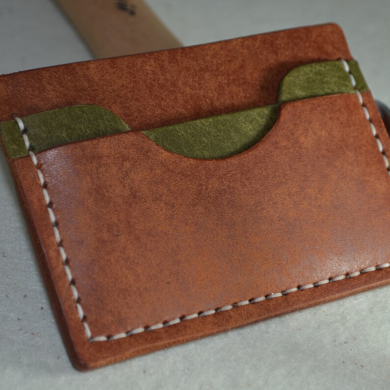 May include: A handmade leather card wallet in shades of brown and olive green. The wallet features a contrasting olive green pocket and is stitched with white thread. The leather has a textured appearance.