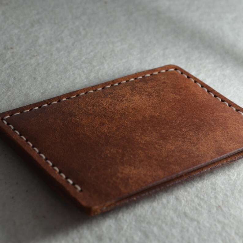 May include: A rectangular, brown leather card holder with white stitching around the edges. The leather has a distressed appearance, with variations in color and texture. The card holder is likely designed to hold credit cards or business cards.