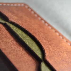 May include: Close-up of a handcrafted leather wallet. The wallet features a rich brown exterior with a contrasting olive green interior. White stitching accents the edges, highlighting the craftsmanship. The leather has a textured appearance.