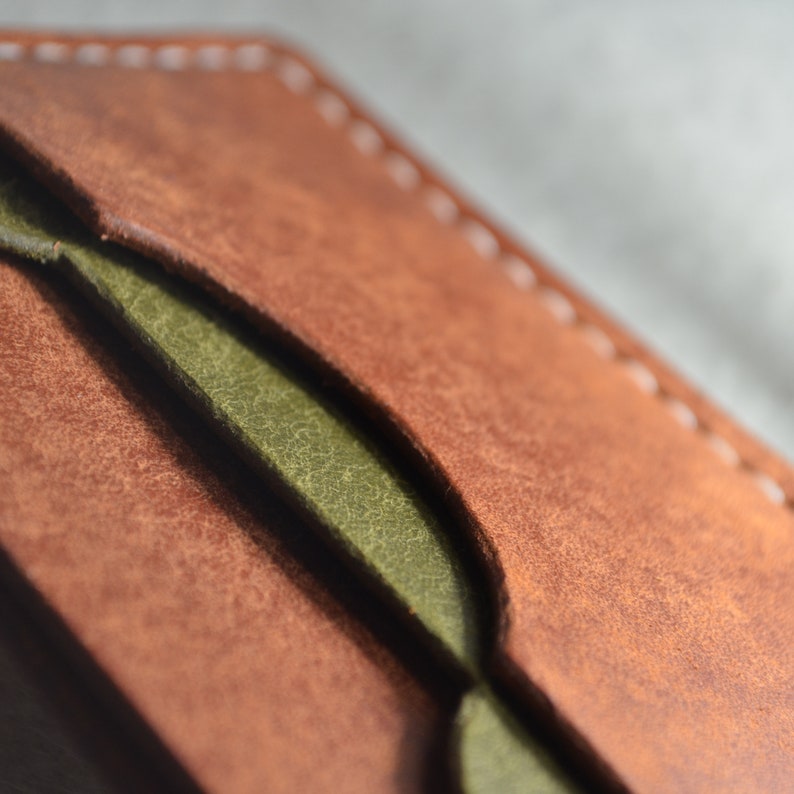 May include: Close-up of a handcrafted leather wallet. The wallet features a rich brown exterior with a contrasting olive green interior. White stitching accents the edges, highlighting the craftsmanship. The leather has a textured appearance.