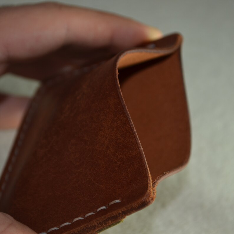 May include: A brown leather wallet with white stitching. The wallet is open, revealing a lighter brown interior. The leather has a textured appearance. The wallet is held in a person's hand.