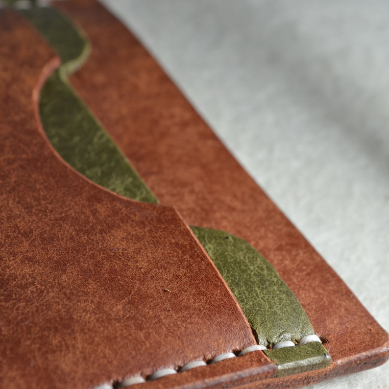 May include: Close-up of a brown leather wallet with olive green accents. The wallet features a bi-fold design with visible stitching and curved cutouts. The leather has a textured appearance.