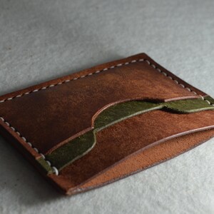 May include: A handcrafted leather card holder in shades of brown and olive green. The wallet features a textured, distressed finish and is stitched with white thread. The design includes a decorative green accent.